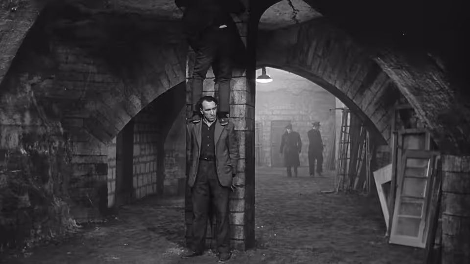 A man stands beneath stone arches in a prison cellar as two figures approach