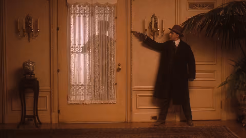 Vito Corleone (Robert De Niro) aiming a pistol at a lace-curtained door