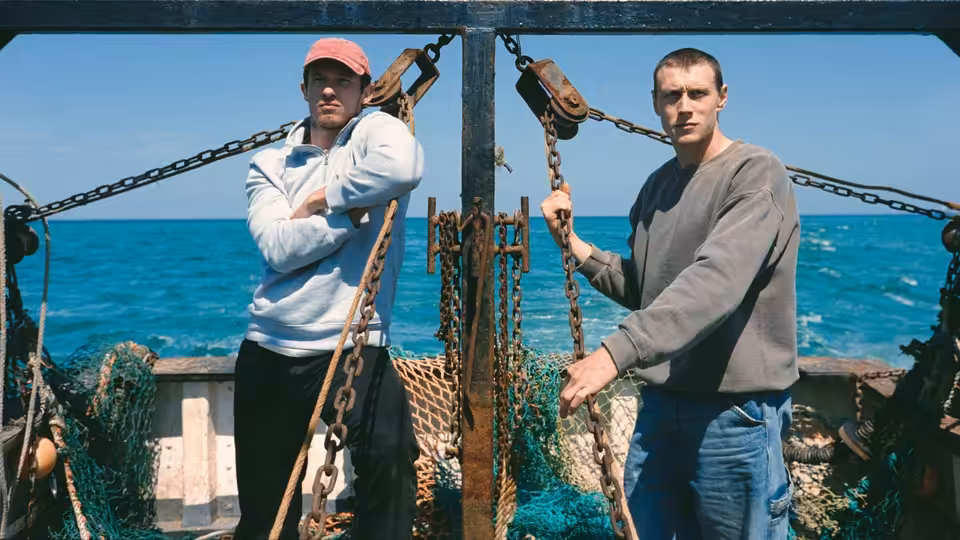 Two men stand on a fishing boat deck by the sea, one with arms crossed