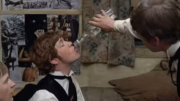 student drinking directly from a glass bottle held by another in a bedroom