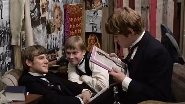 three students in uniform examining a folder together in a cluttered dormitory room