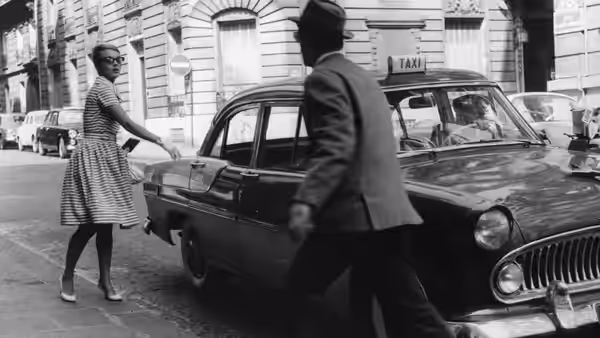 A woman reaches for a taxi door on a city street as a man approaches