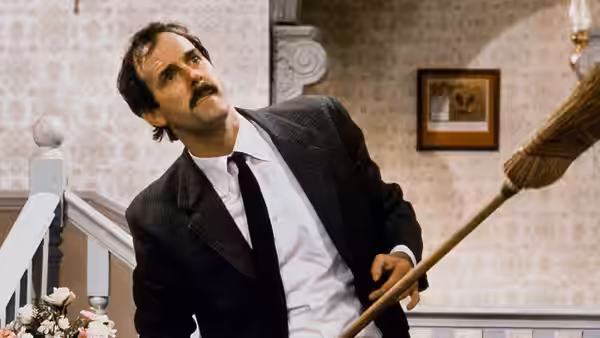 Basil Fawlty (John Cleese) holds a broom and looks upwards on a staircase landing