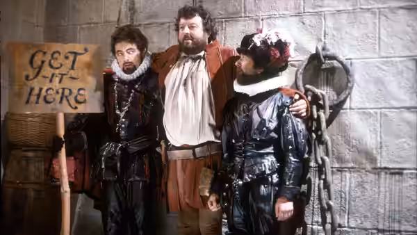 Edmund Blackadder (Rowan Atkinson) standing with two companions beside a stone wall