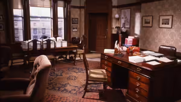 An empty office with wooden desks, chairs, papers and tall windows