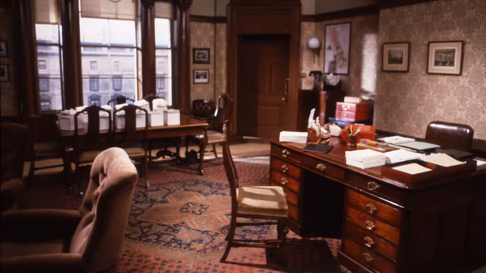 An empty office with wooden desks, chairs, papers and tall windows