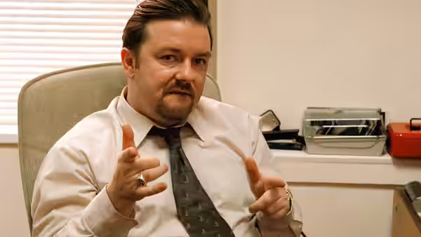 David Brent (Ricky Gervais) gesturing with both hands while seated in an office chair