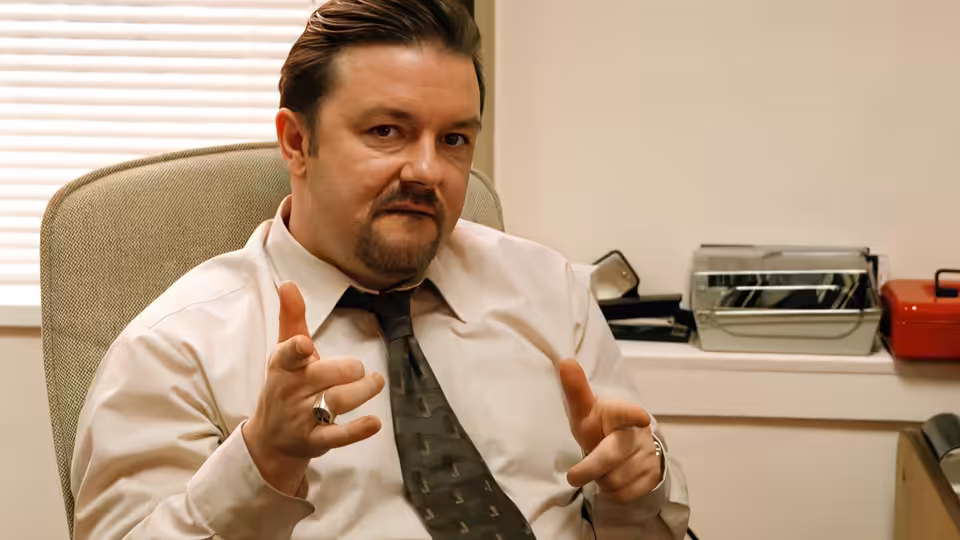 David Brent (Ricky Gervais) gesturing with both hands while seated in an office chair