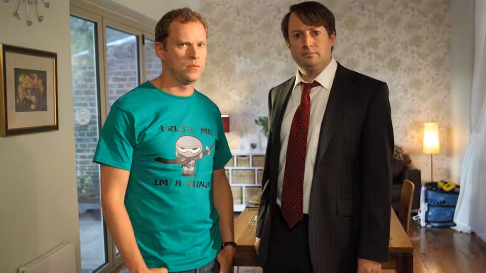 Mark Corrigan (David Mitchell) and Jeremy Usborne (Robert Webb) standing in a living room