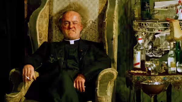 Father Jack Hackett (Frank Kelly) seated in an armchair beside a table of bottles