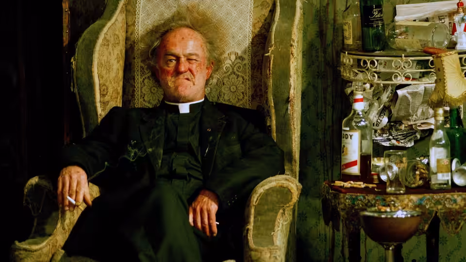 Father Jack Hackett (Frank Kelly) seated in an armchair beside a table of bottles