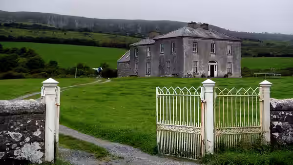 a large grey stone house in a green field with a white gate in front