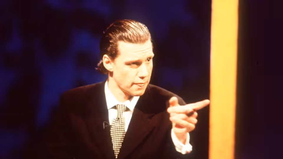 Chris Morris in a dark suit pointing forward with his right hand