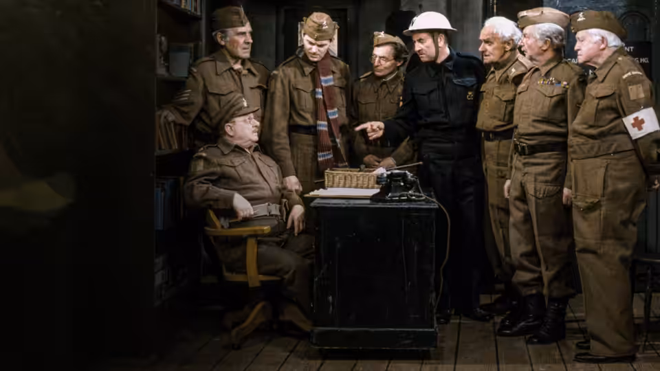 Captain Mainwaring seated at a desk with typewriter as soldiers gather around