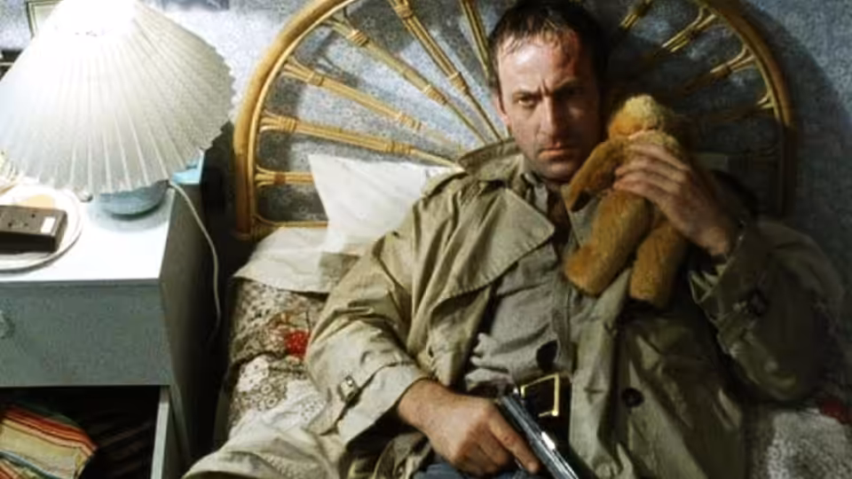Ronald Craven (Bob Peck) lies on a bed holding a teddy bear and pistol
