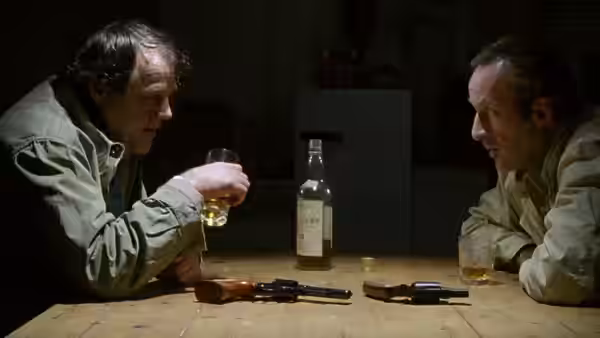 Ronald Craven (Bob Peck) and Darius Jedburgh (Joe Don Baker) drink at a table with guns