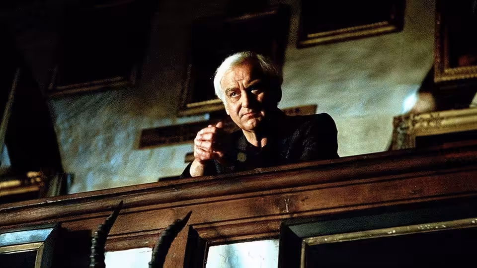 An older man leans forward over a wooden railing in a formal hall