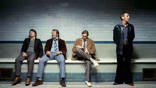 Sam Tyler (John Simm) standing while Gene Hunt (Philip Glenister) and two men sit in a subway