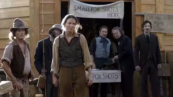 A woman stands before a wooden building with a smallpox shots sign