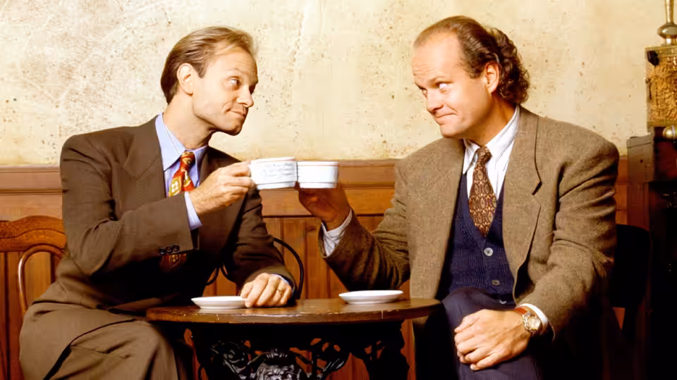Frasier (Kelsey Grammer) and Niles (David Hyde Pierce) toasting with cups at a table