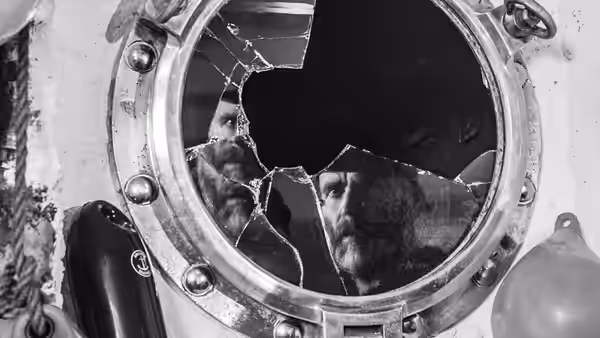 A bearded man looks through the cracked glass of a porthole