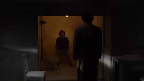 A man stands in a dark washroom doorway facing a seated figure inside