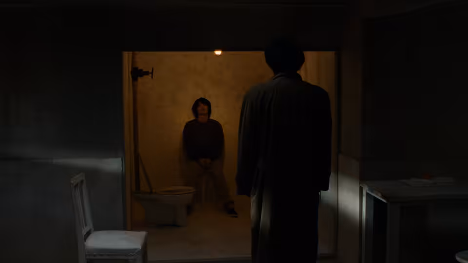 A man stands in a dark washroom doorway facing a seated figure inside