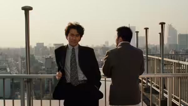 Two suited men talk on a rooftop overlooking the city