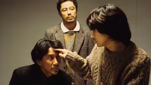 A long-haired man touches another man's forehead as a third watches