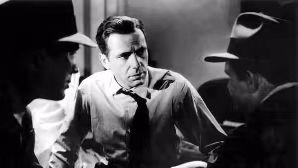 Samuel Spade (Humphrey Bogart) sits at a table facing two men in hats during an interrogation