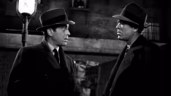 Two men in fedoras stand facing each other on a dimly lit city street at night