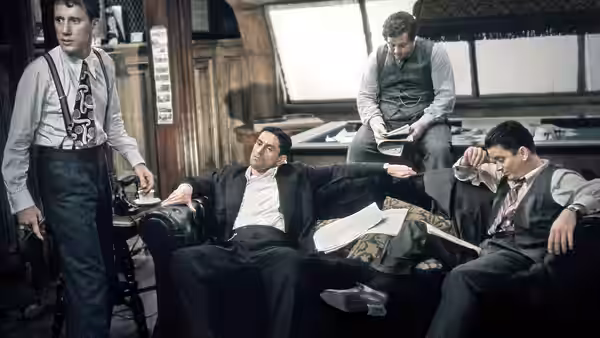Robert De Niro reclining on a couch in an office with other men standing nearby
