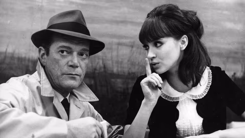 Lemmy Caution (Eddie Constantine) and Natacha von Braun (Anna Karina) sit together at a table