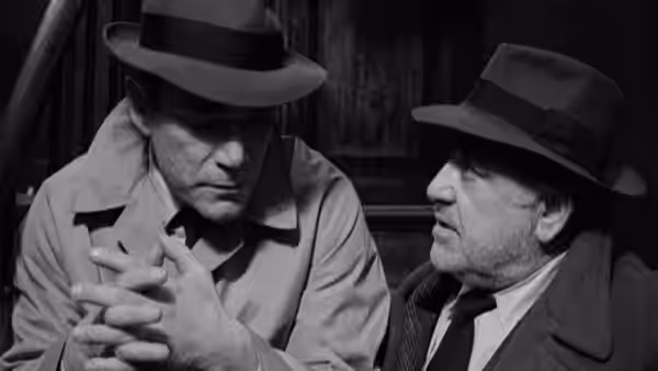 Two fedora-wearing men sit facing each other in a dark room