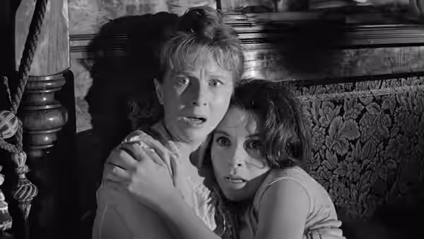 A frightened woman clutches another woman beside patterned wallpaper