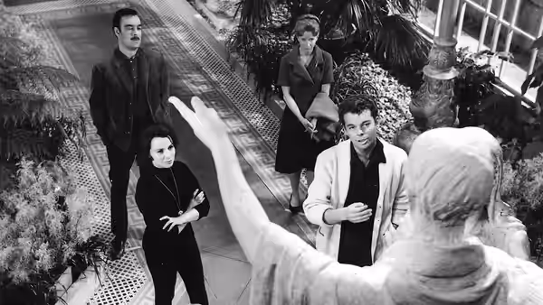 A woman raises her arm toward four visitors gathered in the mansion hall