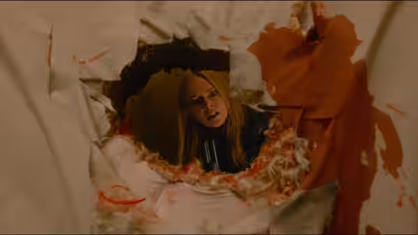 A blonde woman peers through a jagged hole in a wall