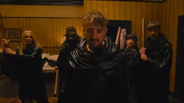 A blond man stands among black-robed figures in a wood-paneled room