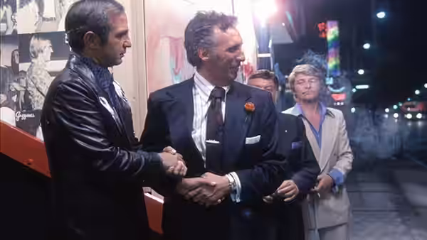 Cosmo Vittelli (Ben Gazzara) shakes hands with a man outside a theatre as Mort Weil (Seymour Cassel) watches