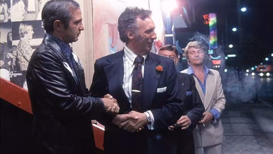 Cosmo Vittelli (Ben Gazzara) shakes hands with a man outside a theatre as Mort Weil (Seymour Cassel) watches