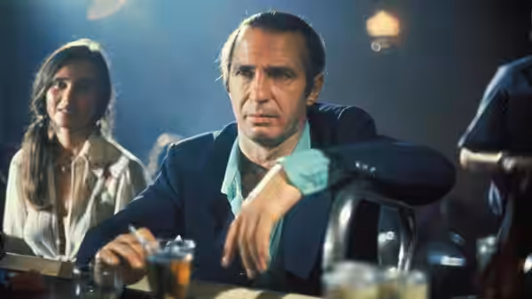Cosmo Vittelli (Ben Gazzara) sits at a table beside a woman, holding a drink