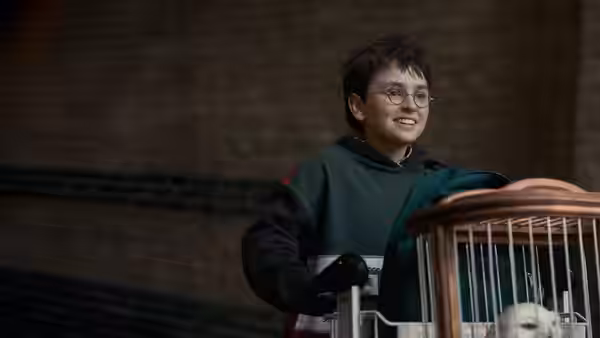 Harry Potter smiles beside a small owl cage in a dim room