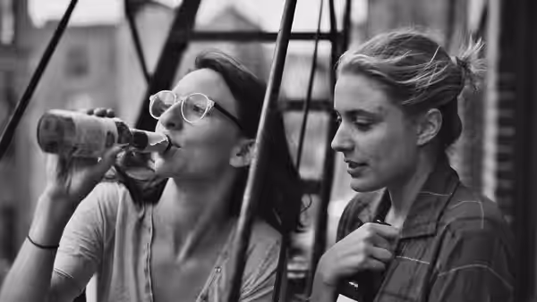 Sophie (Mickey Sumner) drinking from a bottle beside Frances (Greta Gerwig)