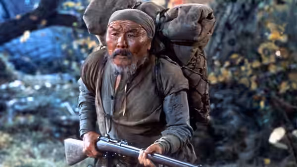 Dersu Uzala (Maksim Munzuk) carries a rifle and a large pack through the forest