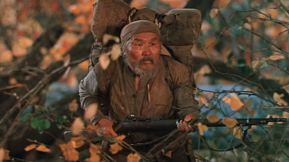 Dersu Uzala (Maksim Munzuk) crouches among autumn leaves, holding a rifle