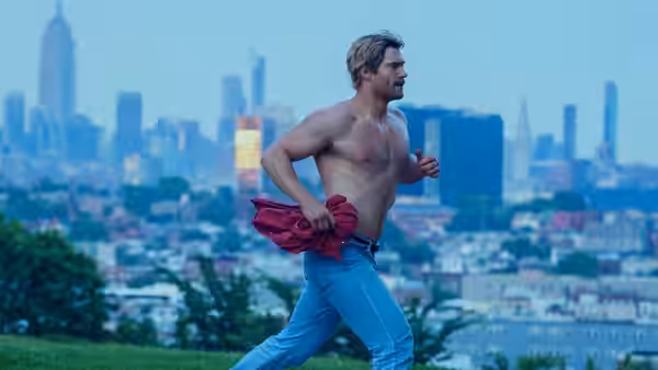 John Tuggle (David Corenswet) runs shirtless along a city waterfront, clutching a red shirt