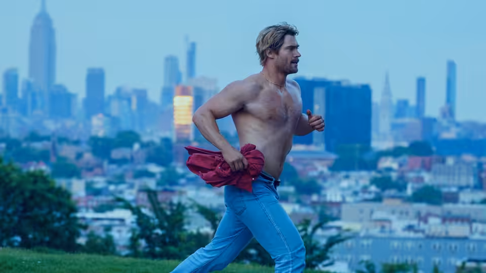 John Tuggle (David Corenswet) runs shirtless along a city waterfront, clutching a red shirt
