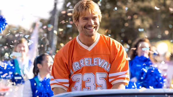 John Tuggle (David Corenswet) smiles in an orange football jersey before a cheering crowd