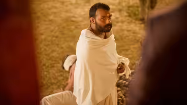 A bearded man in a white wrap stands with a yellow tilak on his forehead