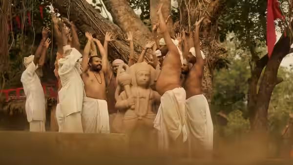 Men in white dhotis raise their arms around a large Hanuman statue beneath a tree